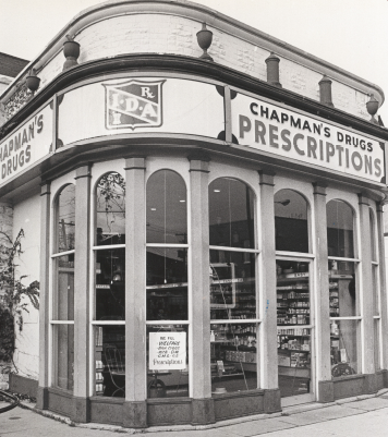 Drug store on a corner with a rounded corner on the building containing tall arched windows. Above this is the drug store sign.